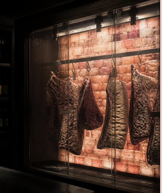 Beef cuts hanging to age behind glass against a wall of glowing Himalayan salt bricks