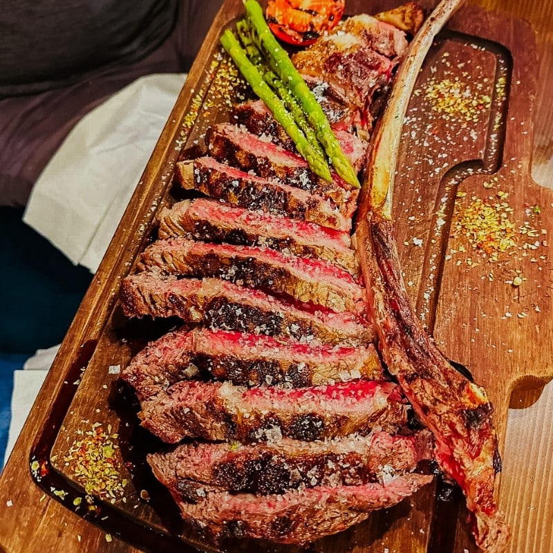 A salt-aged tomahawk steak resting on a charred board beside the charcoal grill